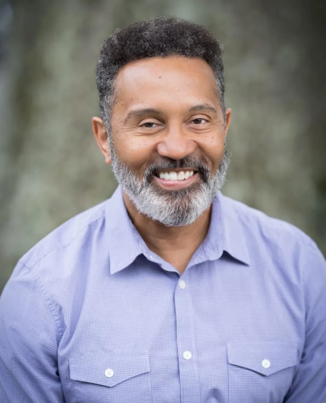 A man sits outside in a button down shirt, smiling.